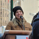 A woman in winter clothing smiles while speaking behind a podium while a crowd looks on.